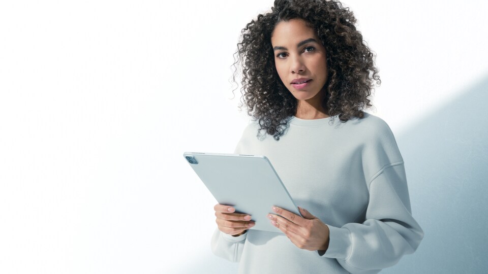 Eine junge Frau mit lockigem Haar, die vor einem weißen Hintergrund in die Kamera blickt, während sie ein Tablet in der Hand hält